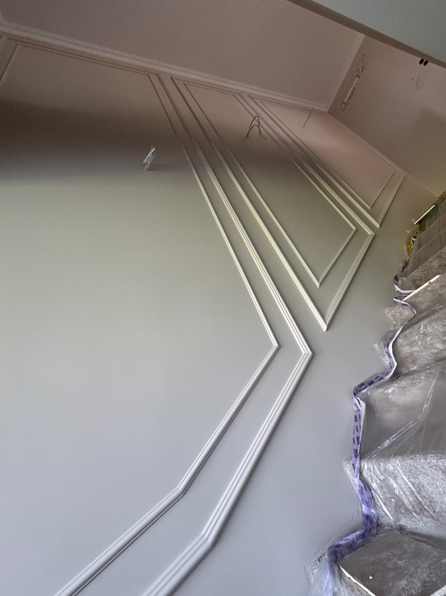 Szara ściana z dekoracyjnymi, białymi listwami w kształcie rombów, częściowo zakryta folią ochronną na schodach. Ujęcie z dołu, podkreślające geometrię wzoru.