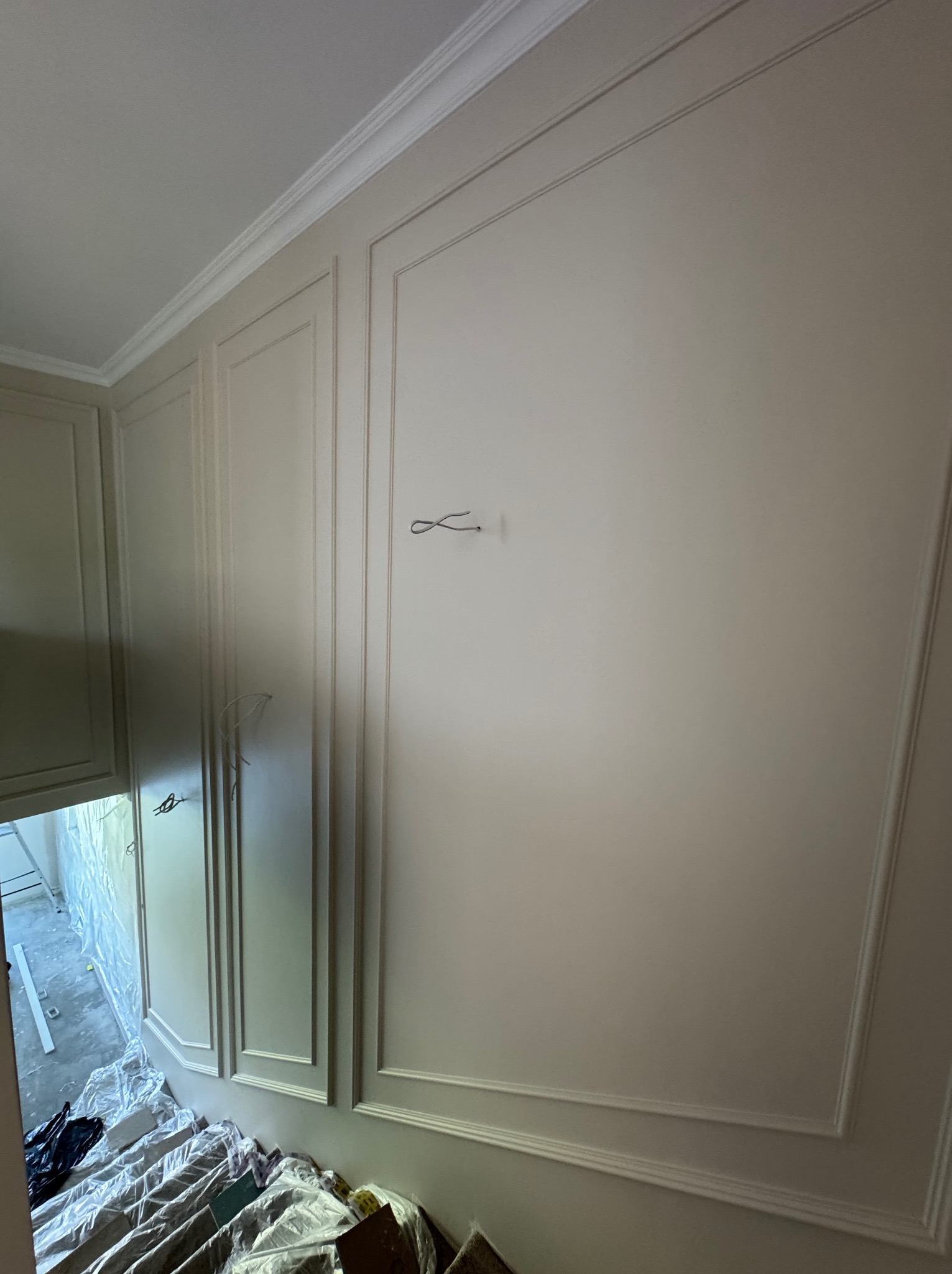 Wykończona ściana z dekoracyjnymi panelami i listwami w jasnym odcieniu. Widoczne przygotowania do dalszych prac wykończeniowych w pomieszczeniu obok.