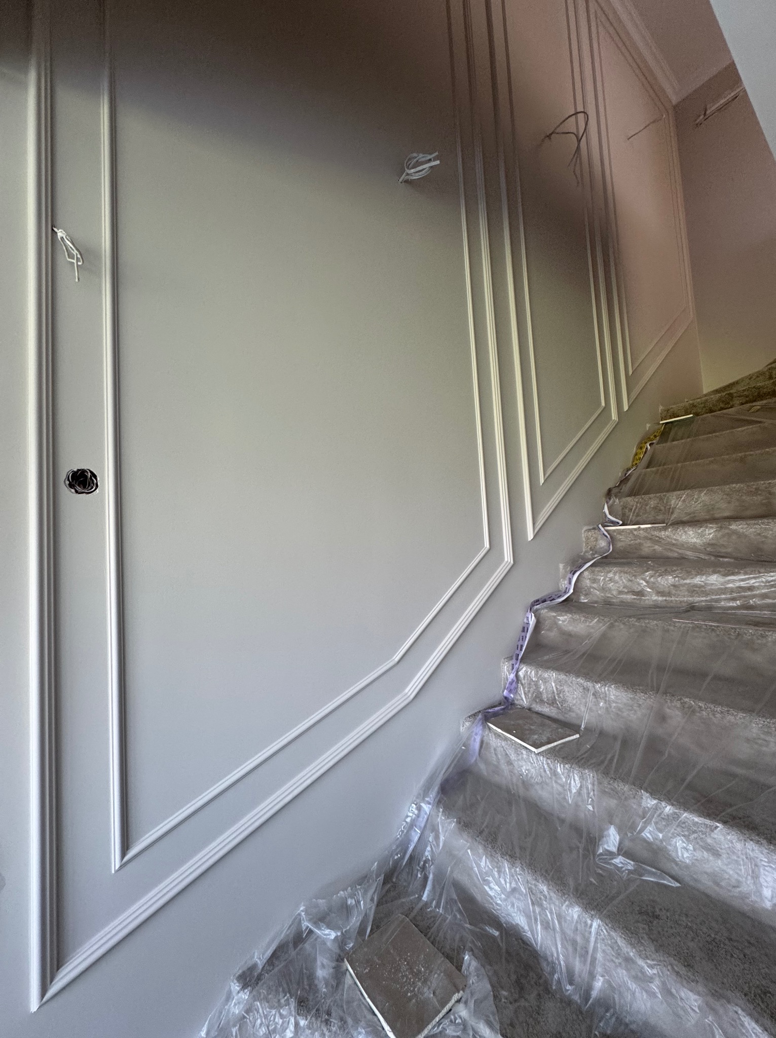 Ściana z dekoracyjnymi listwami sztukateryjnymi w trakcie montażu, widoczne przewody elektryczne i zabezpieczone schody folią. Szare odcienie.