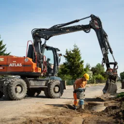 Pracownik w żółtym kasku zagęszcza grunt wokół studzienki rewizyjnej przy użyciu ubijarki stopowej; w tle stoi pomarańczowa koparka kołowa Atlas 140W na drodze z widocznymi drzewami i słupami...