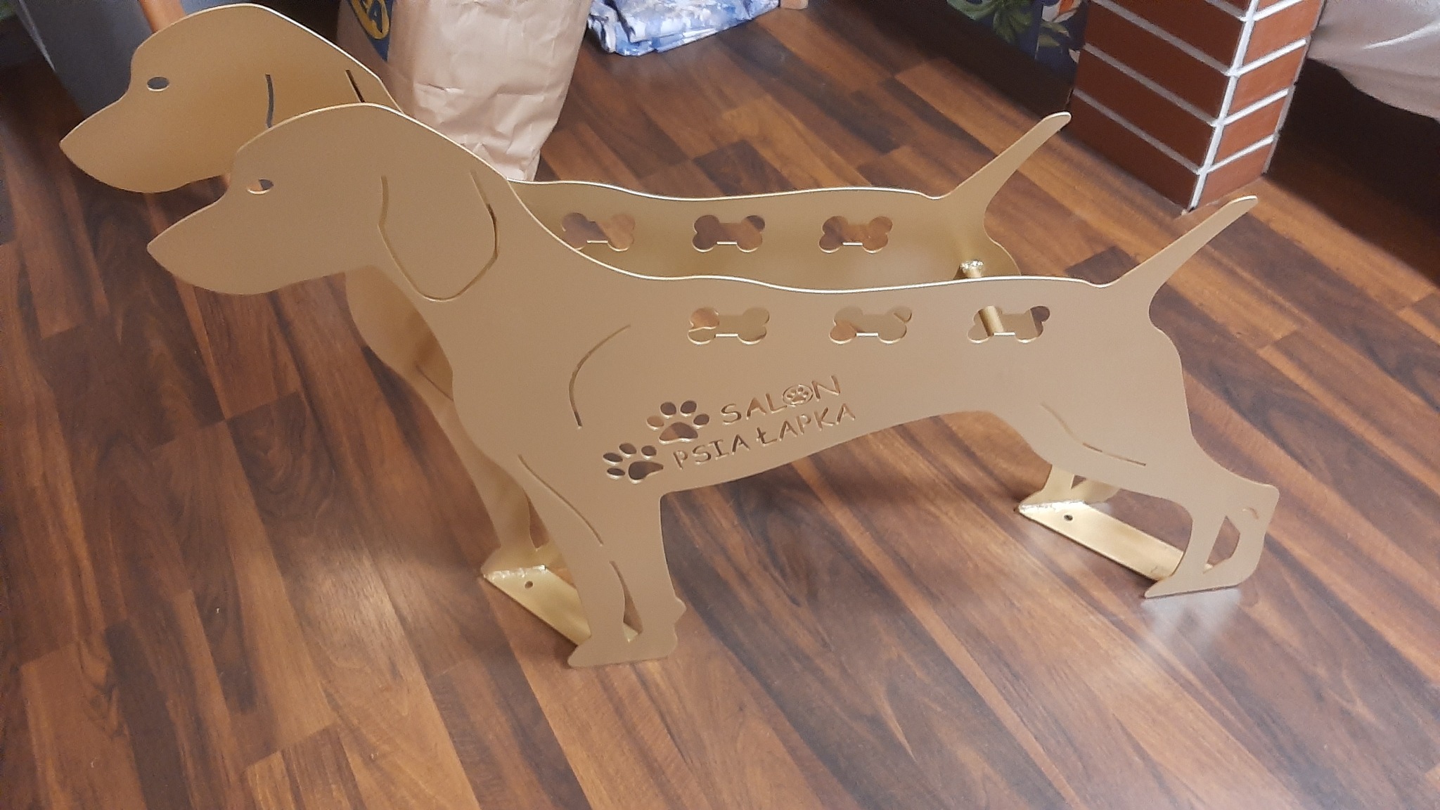 Dwa identyczne, złote, dekoracyjne stojaki w kształcie psów rasy wyżeł, z wyciętymi wzorami kości i napisem 'Salon Psia Łapka', stojące na drewnianej podłodze.