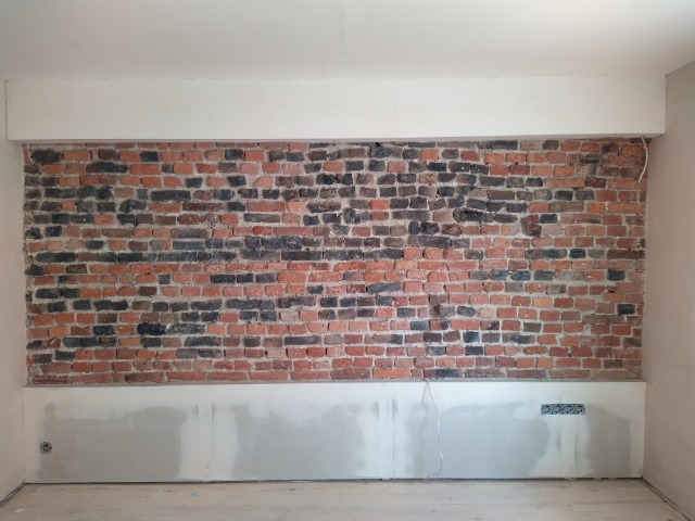 Częściowo odsłonięta, stara ceglana ściana z fragmentami tynku i instalacji elektrycznej, przygotowana do dalszych prac wykończeniowych w pomieszczeniu.