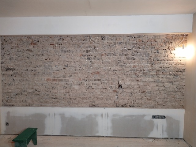 Odsłonięta ceglana ściana w trakcie remontu, z widocznymi śladami po pracach murarskich i elektrycznych, częściowo wykończona gładzią gipsową, oświetlona ciepłym światłem lampy.