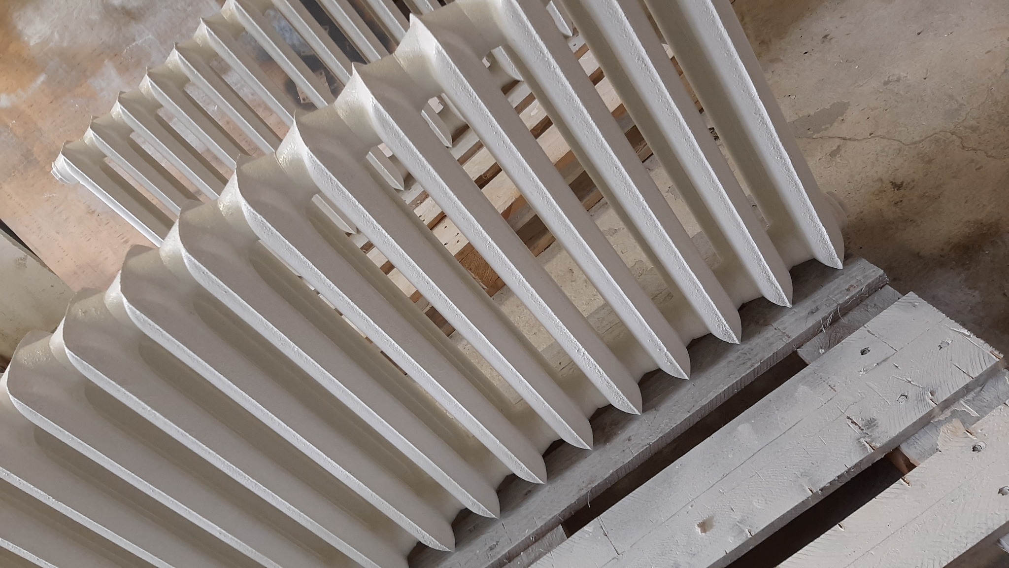 Białe, odnowione żeberkowe grzejniki ułożone na pomalowanej palecie, widoczne ślady po procesie malowania.