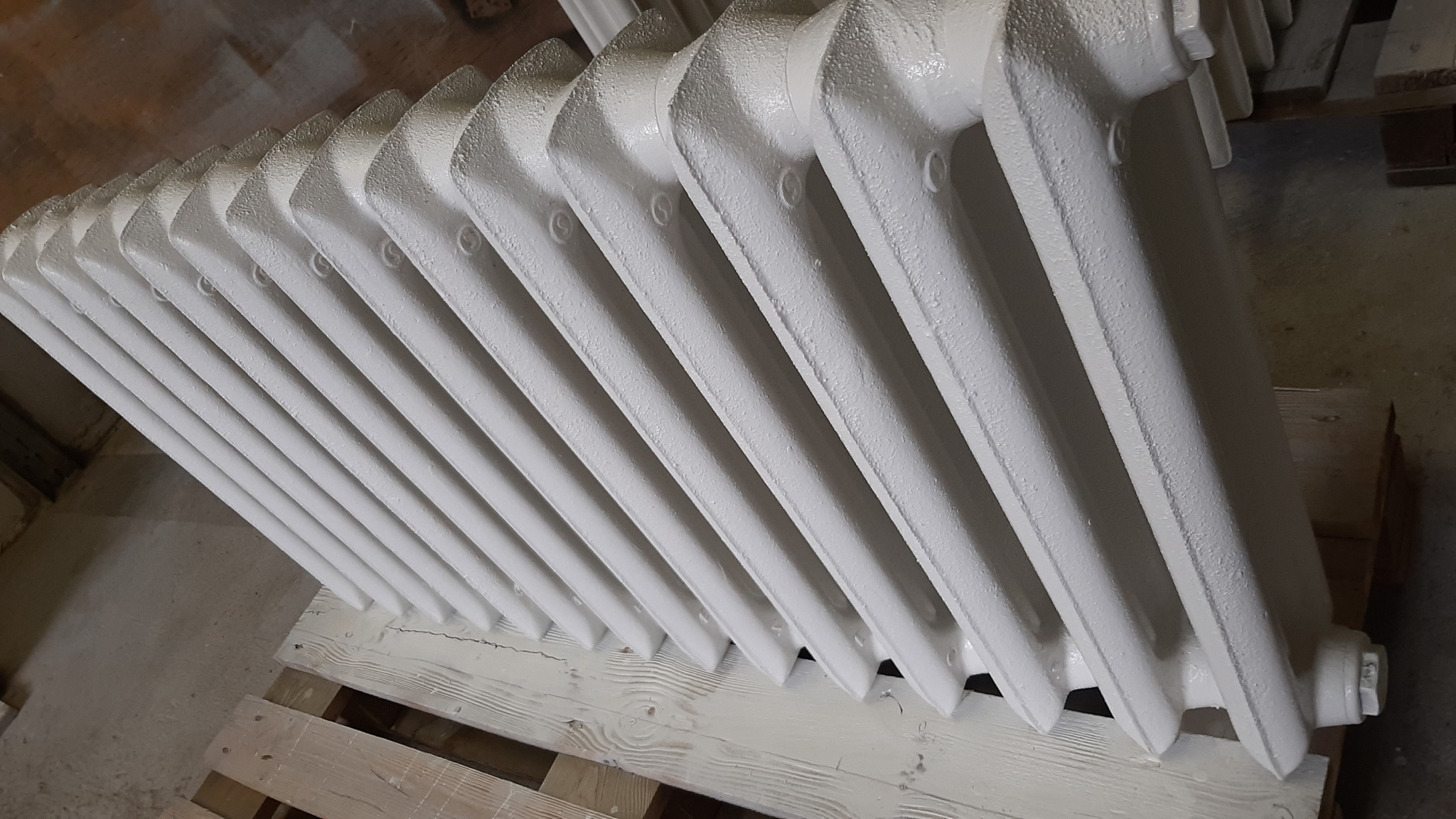 Żeliwny grzejnik po renowacji, pomalowany na biało, leżący na drewnianej palecie.