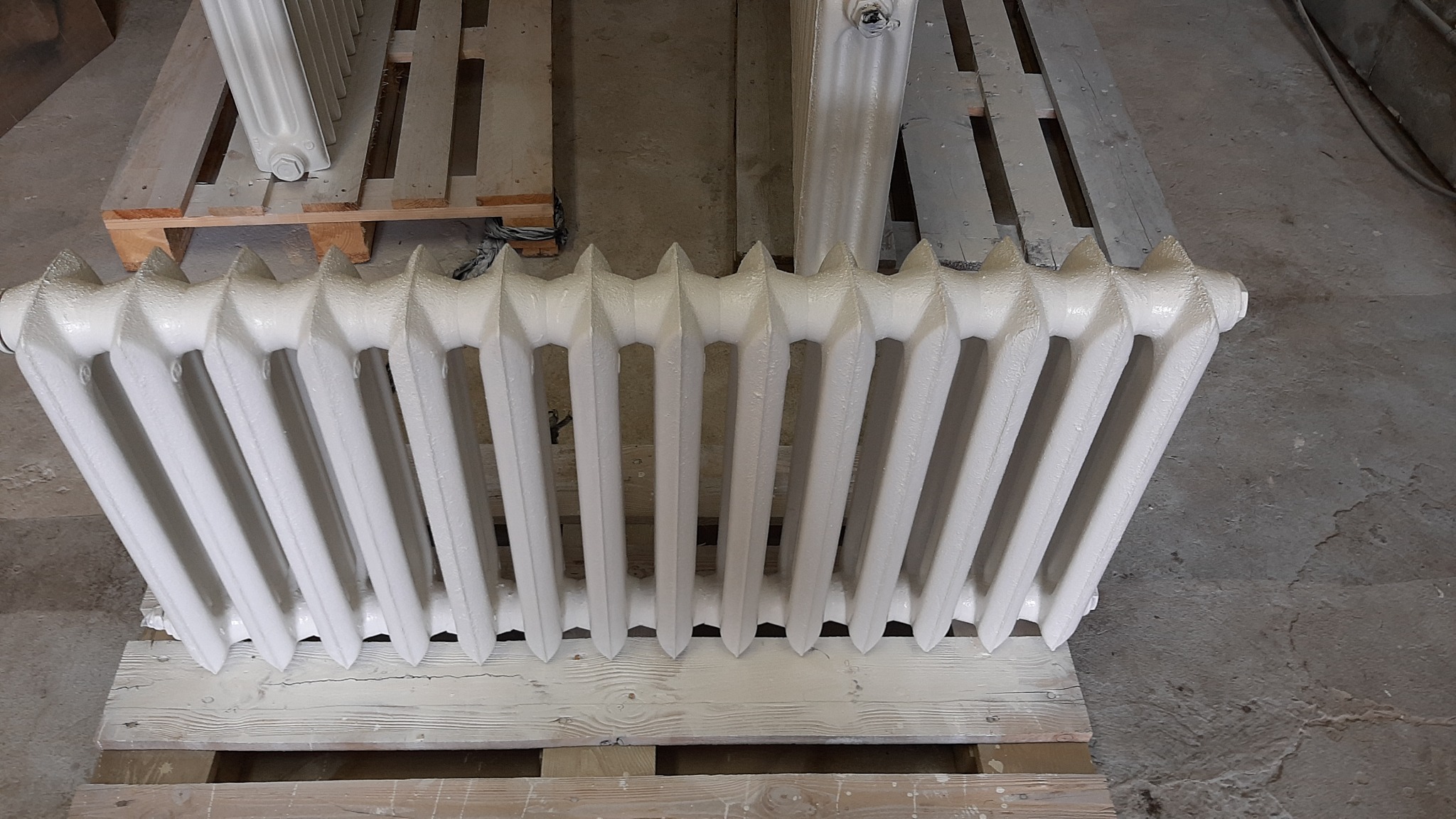 Biały, żeliwny grzejnik po piaskowaniu i malowaniu proszkowym, leżący na drewnianej palecie w szarej hali.