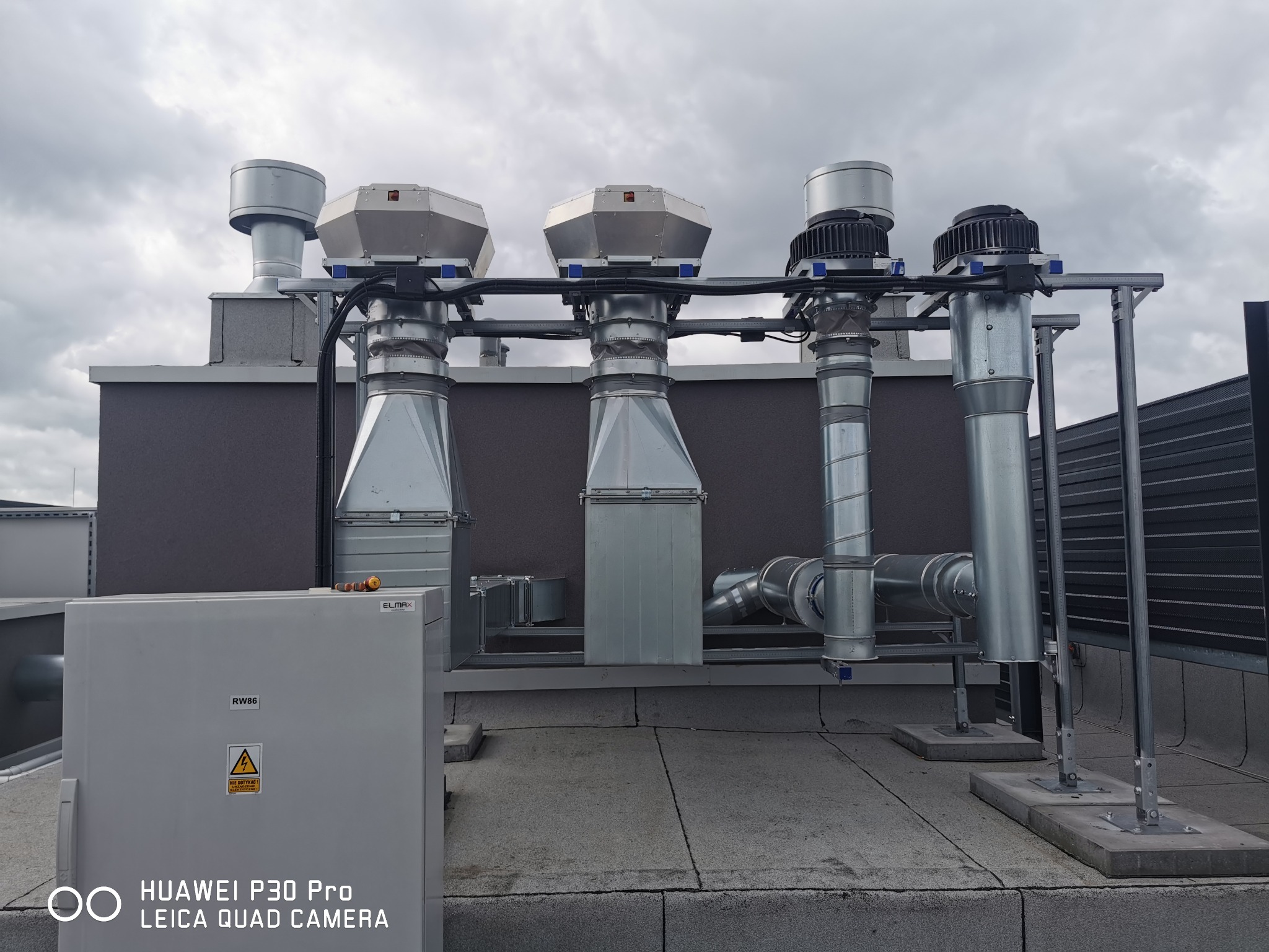 Instalacja wentylacyjna na dachu budynku z czterema pionowymi kominami, centralą sterującą ELMAX RW86 i pochmurnym niebem w tle.