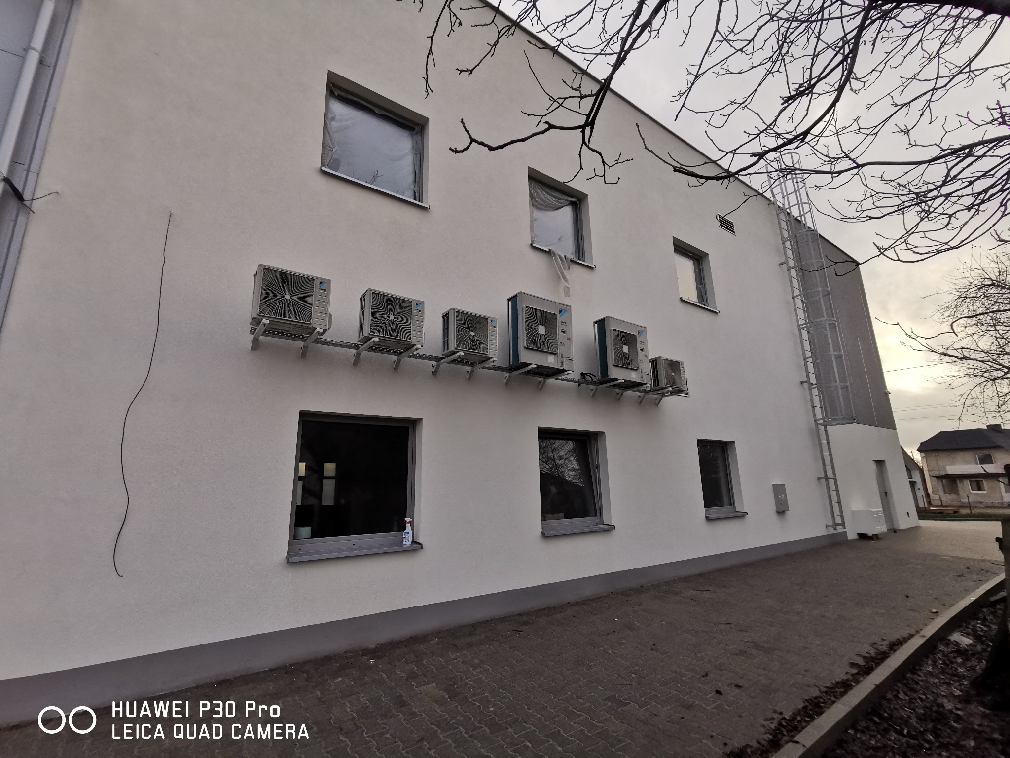 Elewacja budynku z zamontowanymi jednostkami zewnętrznymi klimatyzacji na wspólnym stelażu, okna zabezpieczone folią, widoczna drabina ewakuacyjna i butelka płynu do czyszczenia na parapecie.