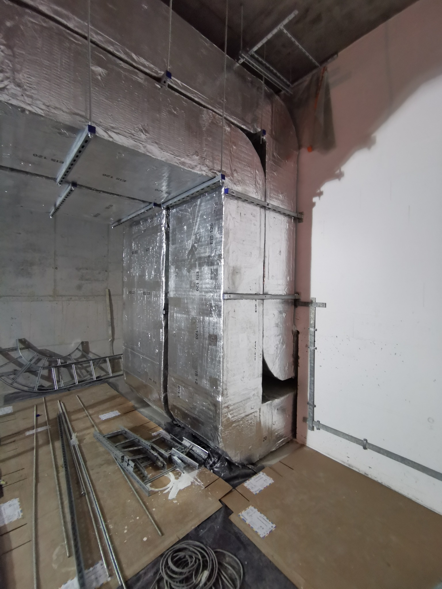 Montaż izolowanej instalacji wentylacyjnej w surowym wnętrzu, widoczne elementy mocujące, drabina i kable na podłodze zabezpieczonej kartonem.