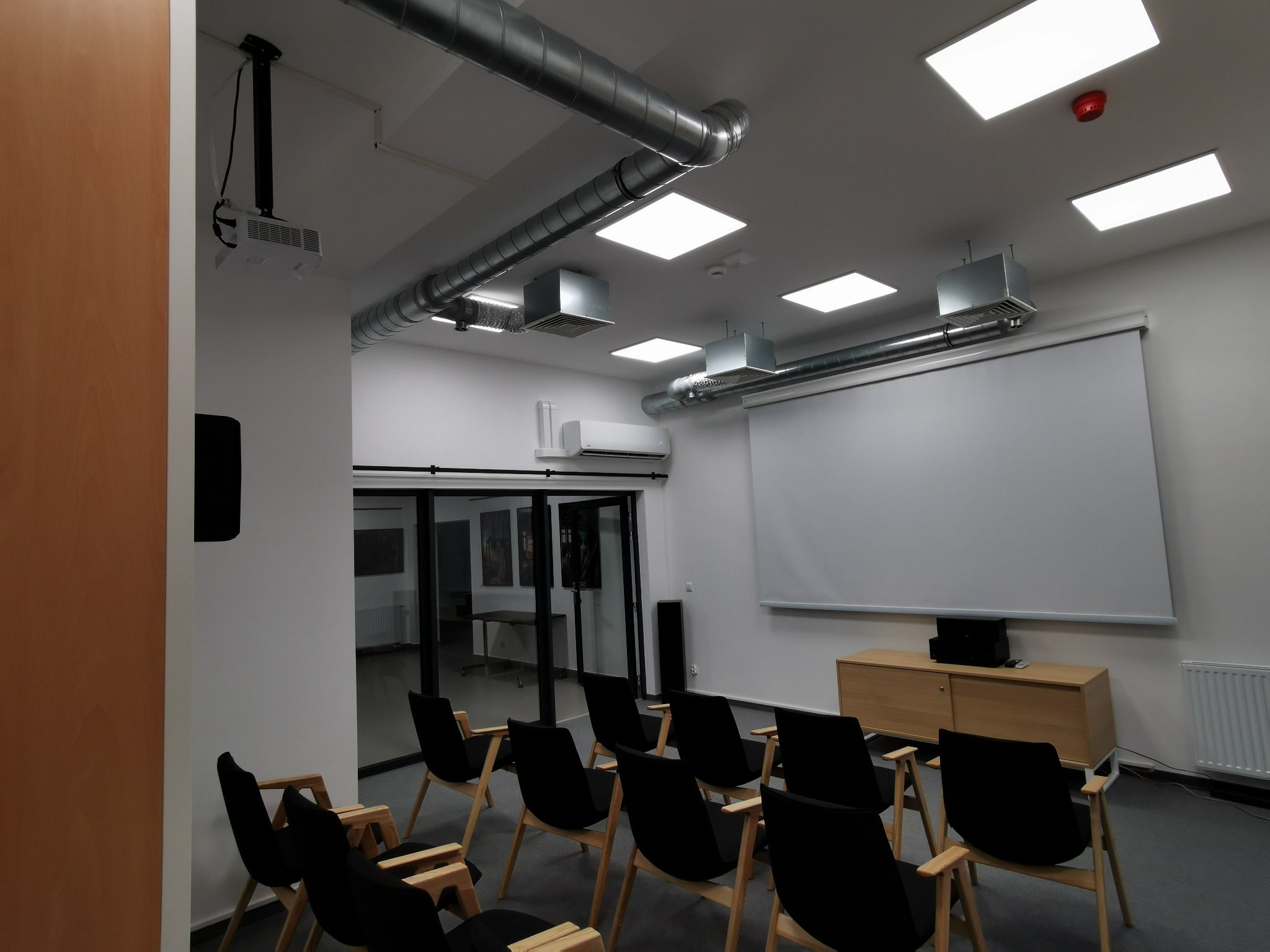 Sala konferencyjna z zamontowaną instalacją wentylacyjną, klimatyzatorem i projektorem, widoczne metalowe rury wentylacyjne pod sufitem, kratki wentylacyjne oraz klimatyzator ścienny.