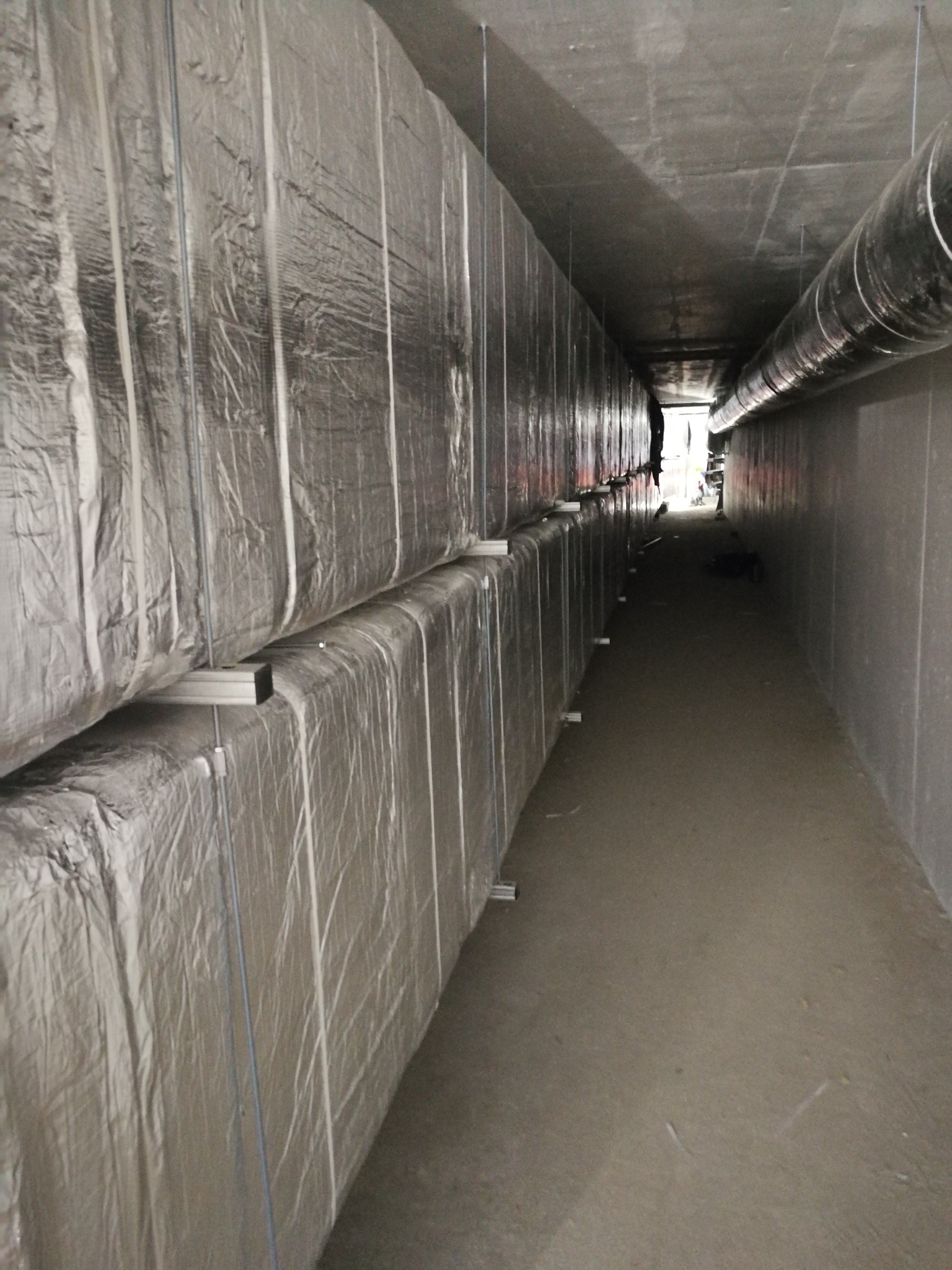 Długi korytarz z izolowanymi przewodami wentylacyjnymi pod sufitem, widoczne metalowe wsporniki i srebrna folia izolacyjna.