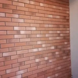 Ściana wyłożona cegłą klinkierową w odcieniach czerwieni i brązu, z widoczną fakturą i fugami, oświetlona z boku, tworząca efekt trójwymiarowości.