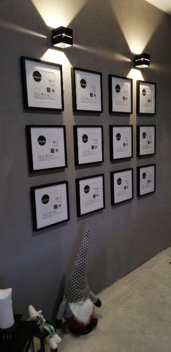 Ściana pomalowana na szaro z symetrycznie rozmieszczonymi dwunastoma czarnymi ramkami z białymi wkładami, oświetlona czarnymi, kwadratowymi kinkietami dającymi podwójne strumienie światła;...