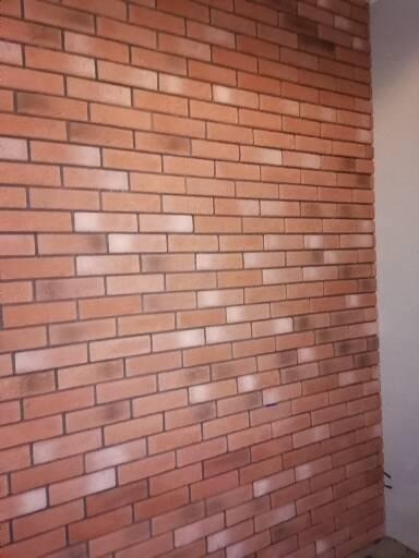 Ściana wyłożona cegłą klinkierową w odcieniach czerwieni i brązu, z widoczną fakturą i fugami, oświetlona z boku, tworząca efekt trójwymiarowości.