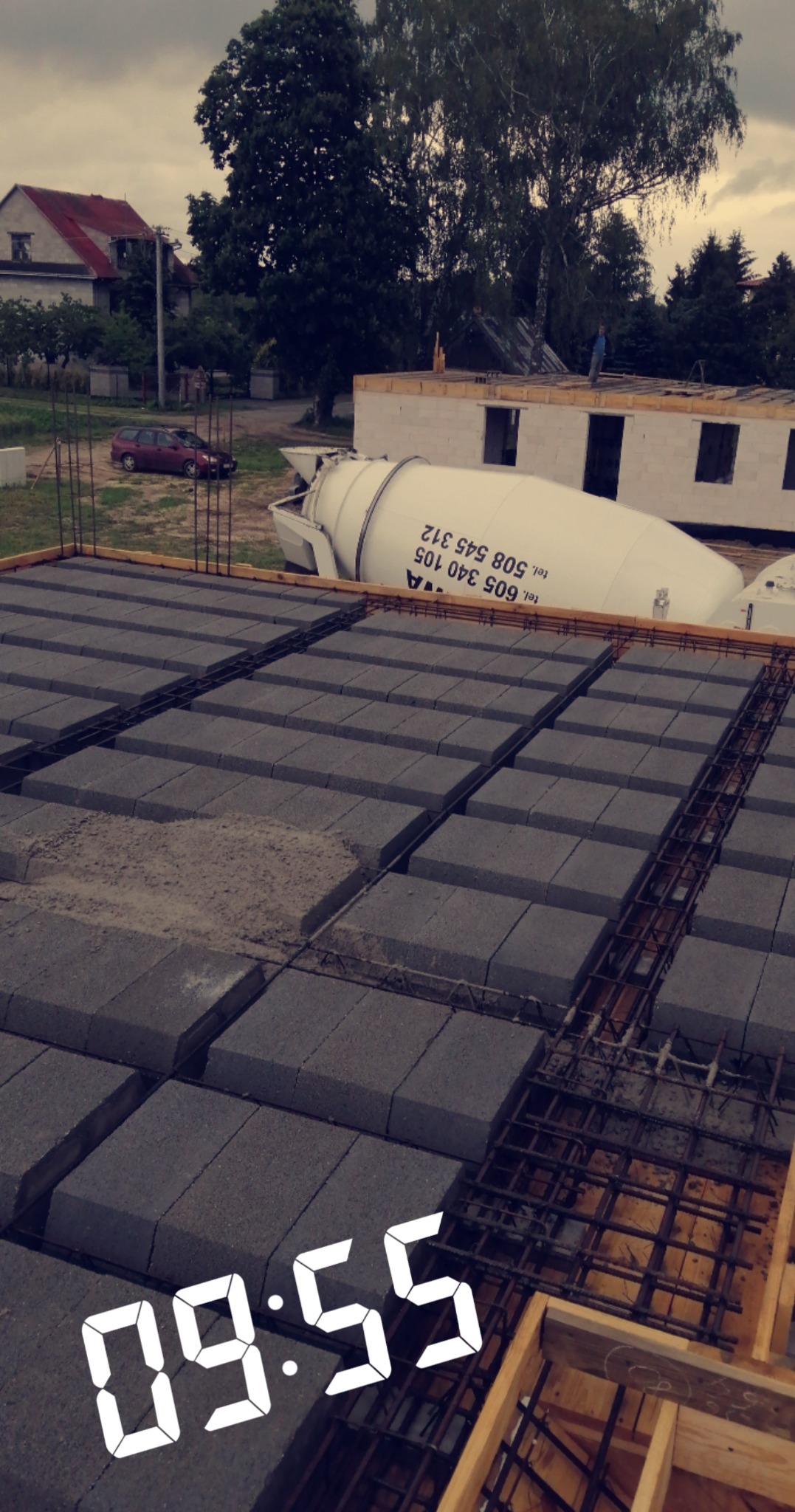 Budowa domu murowanego: widok na strop Teriva z betonowym mikserem w tle, stalowe zbrojenia wystają ponad pustaki, na drugim planie budynek w trakcie wznoszenia ścian.