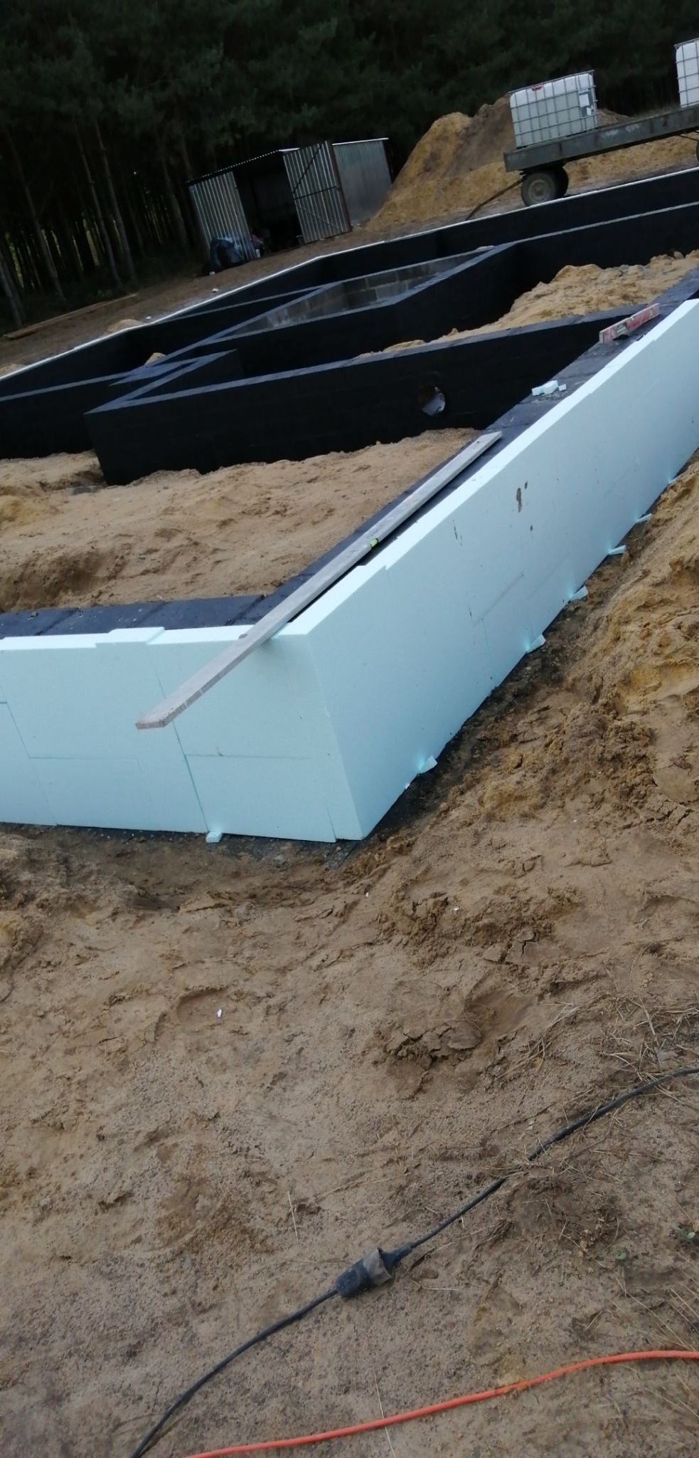 Budowa fundamentów domu: białe płyty izolacyjne ułożone w wykopie, zabezpieczone czarną folią, widoczny poziomica i piasek.