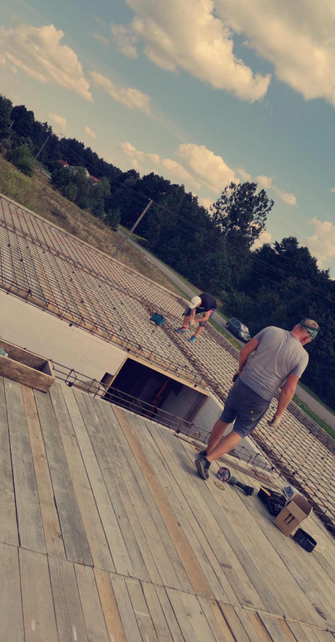 Dwóch pracowników na budowie domu murowanego, układających zbrojenie na styropianie, widok z góry na drewniany pomost, w tle błękitne niebo z chmurami i zieleń drzew.