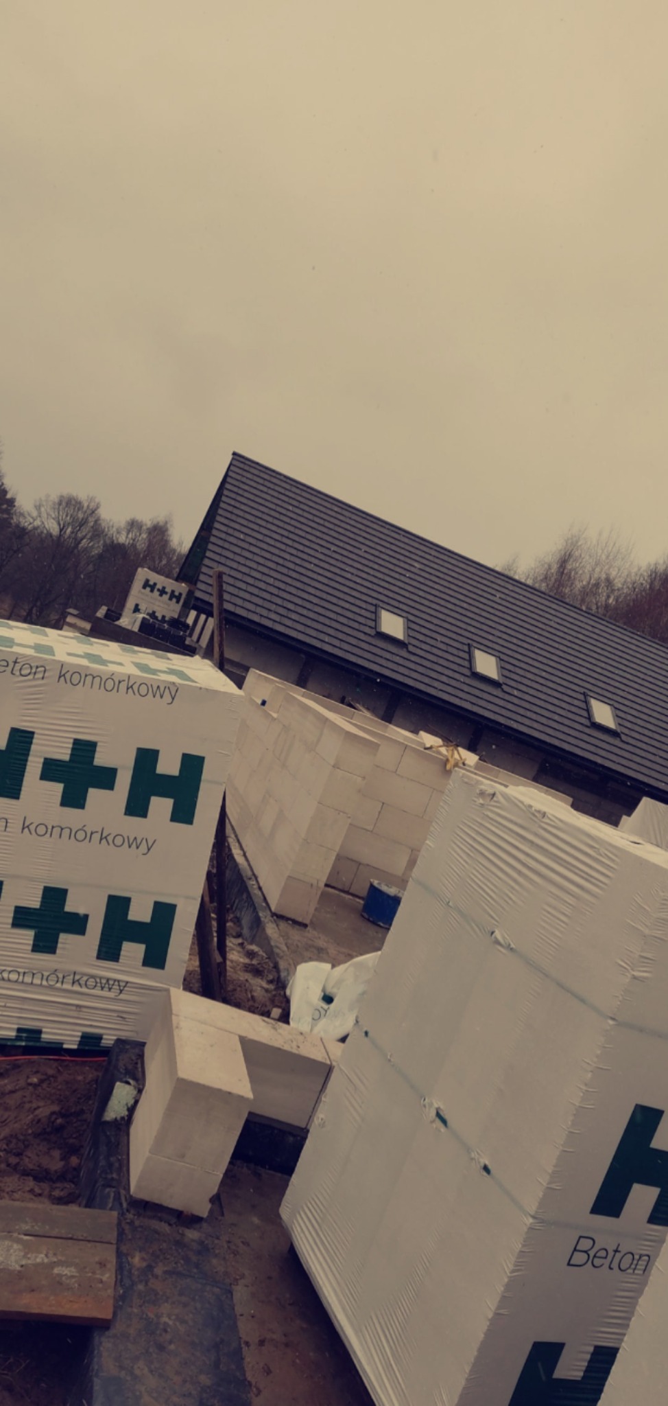 Paczki betonu komórkowego H+H w folii na budowie domu jednorodzinnego, widoczny fragment fundamentu i ściany, w tle dach z oknami połaciowymi i pochmurne niebo.
