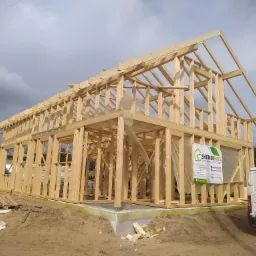 Surowy szkielet domu w technologii szkieletowej, z widocznym fundamentem, paletą budowlaną i samochodem dostawczym firmy Svenskhus z logo i danymi kontaktowymi na banerze i pojeździe.