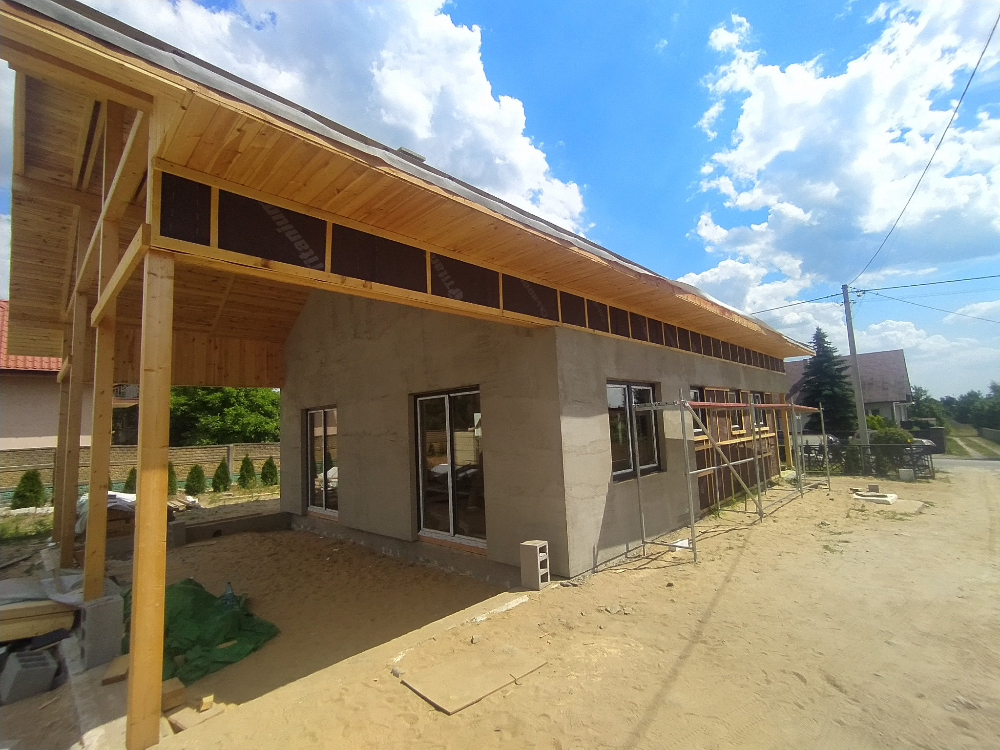 Surowy stan budynku z drewnianą konstrukcją dachu i częściowo otynkowanymi ścianami, widoczne rusztowania i niezabezpieczony teren wokół