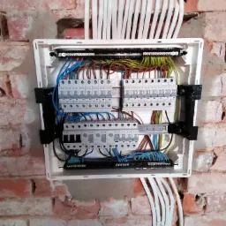 Rozdzielnica elektryczna z zainstalowanymi bezpiecznikami i przewodami w kolorach niebieskim, brązowym, żółto-zielonym, zamontowana na surowej ścianie z czerwonej cegły.