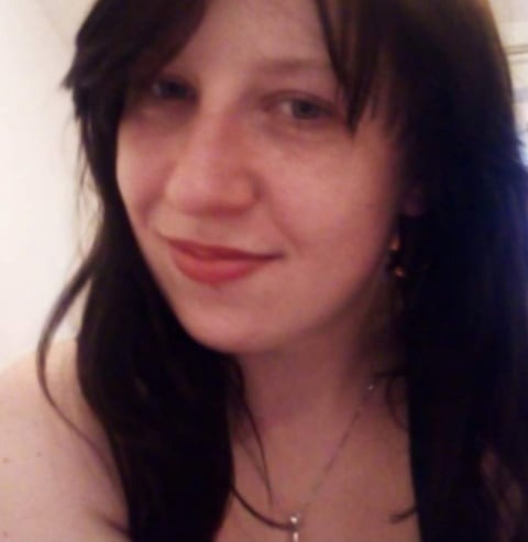 Portret kobiety z ciemnymi włosami, delikatnym uśmiechem i subtelnym makijażem ust, noszącej srebrny naszyjnik z wisiorkiem i małe kolczyki.