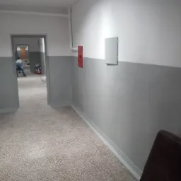 Korytarz w trakcie odświeżania: dolna część ścian pomalowana na szaro, górna na biało, widoczna osoba w oddali przy pracy, na ścianie skrzynka hydrantowa i szafka elektryczna.