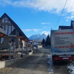 Nowo otynkowany dom w stanie surowym zamkniętym, z widocznymi górami w tle, obok zaparkowana ciężarówka z reklamą usług tynkarskich.