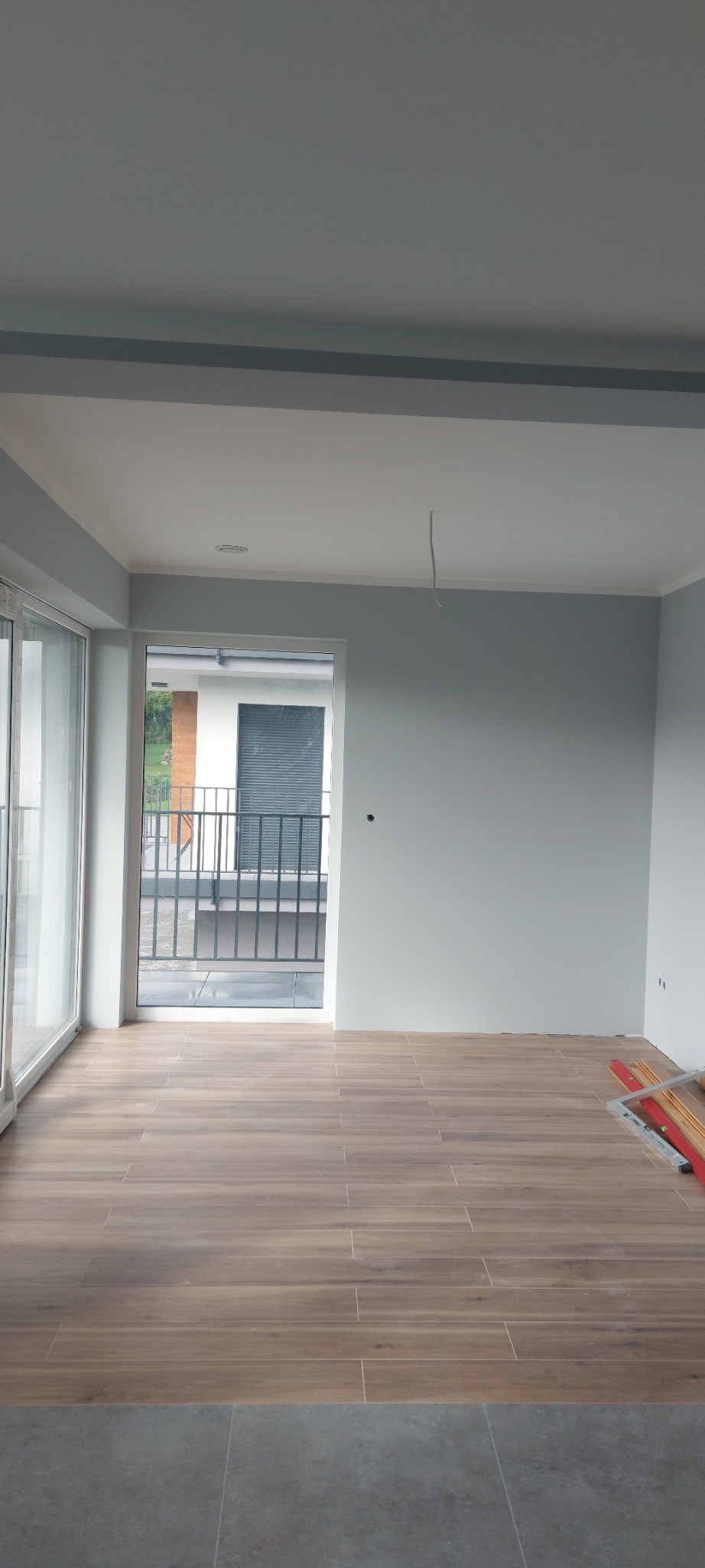 Pomalowane na szaro wnętrze z drewnopodobną podłogą z płytek, widoczne narzędzia malarskie przy ścianie i niedokończone wykończenie podłogi przy progu.