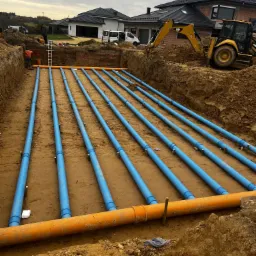 Wykop z ułożonymi niebieskimi rurami instalacyjnymi, widoczny fragment pomarańczowej rury, ziemia, koparka i budynki w tle.