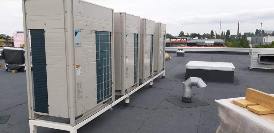 Rząd jednostek zewnętrznych klimatyzacji Daikin VRV IV na dachu budynku komercyjnego, widoczny sklep Bricoman w tle.
