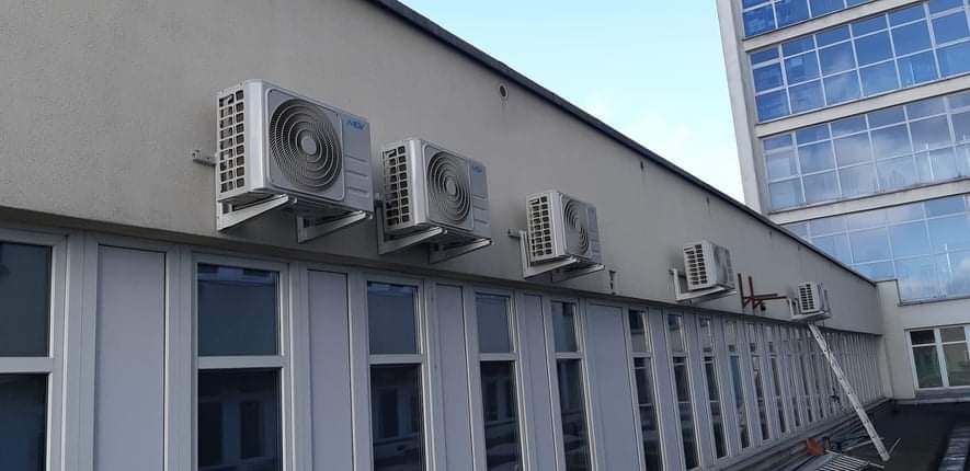 Rząd zewnętrznych jednostek klimatyzacyjnych zamontowanych na ścianie budynku biurowego nad rzędem okien, z rozstawioną drabiną opartą o ścianę.