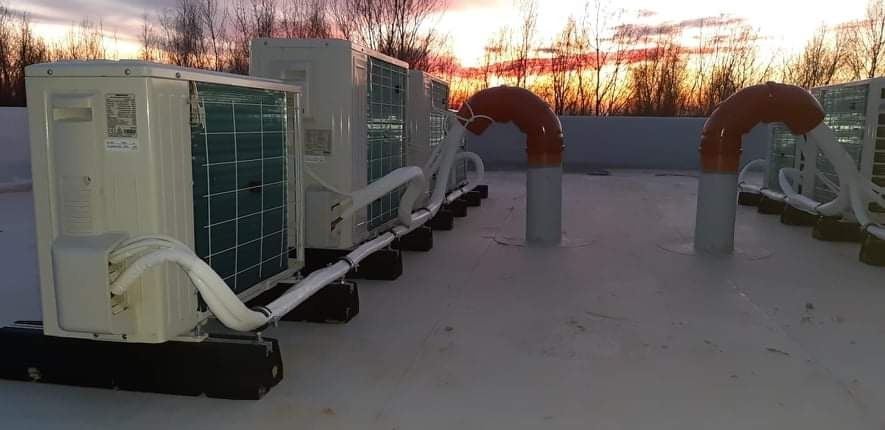 Szereg jednostek zewnętrznych klimatyzacji na dachu budynku komercyjnego, z pomarańczowymi rurami wentylacyjnymi w tle zachodzącego słońca.