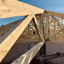 Konstrukcja drewniana dachu w trakcie budowy, widoczne elementy więźby dachowej połączone metalowymi płytkami, na pierwszym planie fragment ściany z bloczków betonu komórkowego, na rusztowaniu piła...