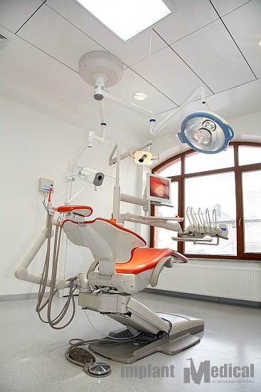 Nowoczesny gabinet dentystyczny z fotelem w odcieniach bieli i pomarańczy, oświetleniem sufitowym i narzędziami na ramieniu, monitor z widokiem zębów, okno z widokiem na miasto.