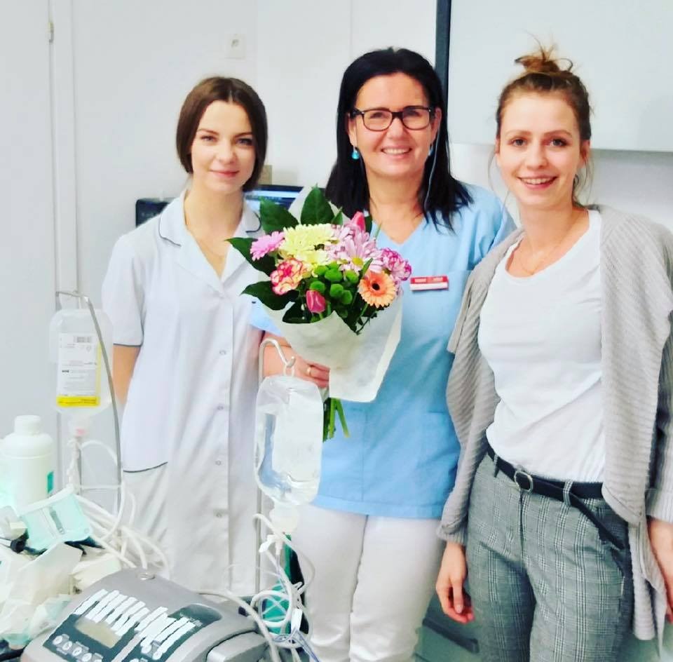 Uśmiechnięte kobiety w placówce medycznej, jedna w stroju medycznym trzyma bukiet kwiatów, widoczne urządzenia medyczne i kroplówki.