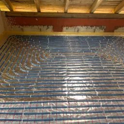 Niebieskie rury ogrzewania podłogowego ułożone spiralnie na folii izolacyjnej z siatką, widoczne drewniane belki stropu i fragment ściany w surowym stanie.