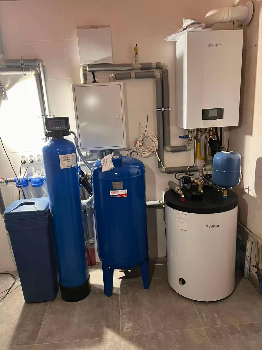 Biały piec gazowy Vaillant z szarymi rurami, obok niebieski zbiornik hydroforowy i zmiękczacz wody w kotłowni z szarą podłogą. Instalacja po montażu.