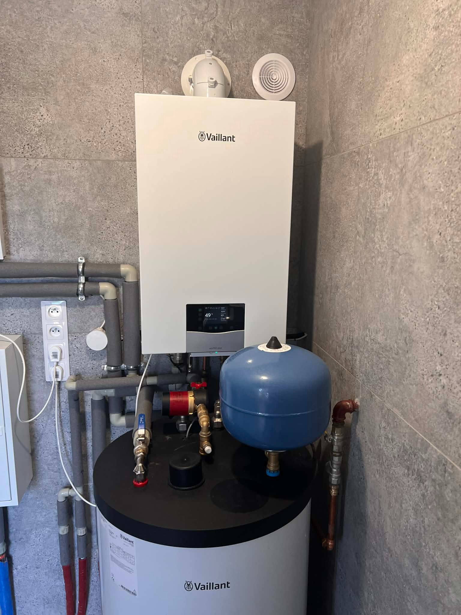 Biały piec gazowy Vaillant zamontowany na tle szarej ściany z płytkami, z widocznymi rurami i elementami instalacji. Ujęcie z bliska, prezentujące estetyczny montaż.