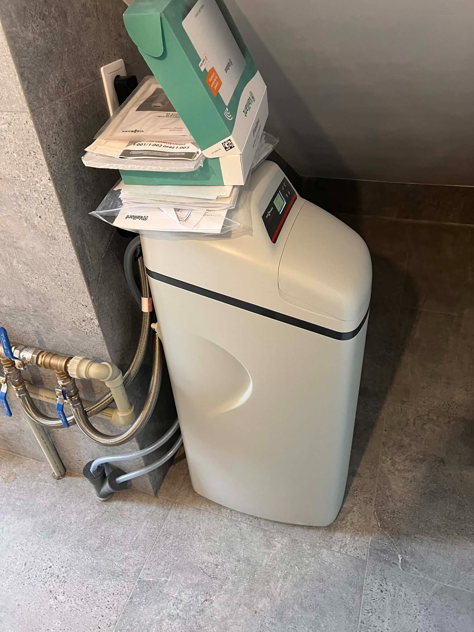 Zamontowany zmiękczacz wody Vaillant z podłączonymi rurami, na którym leżą dokumenty i pudełko. Widoczne przyłącza hydrauliczne i szare płytki na ścianie i podłodze.