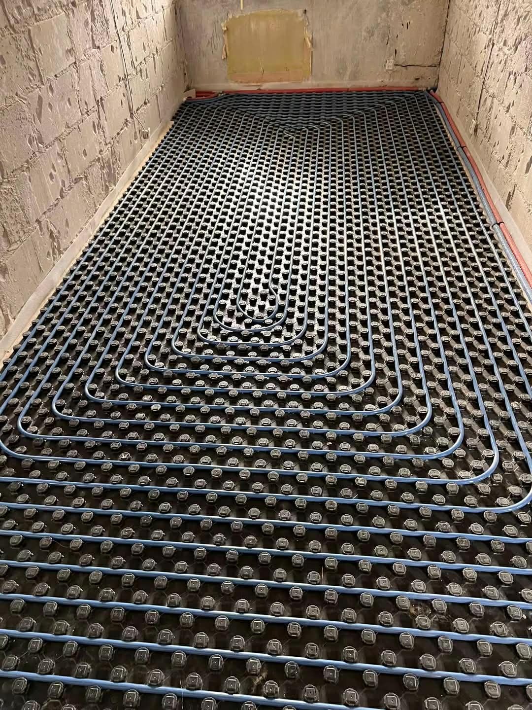 Niebieskie rury ogrzewania podłogowego ułożone na czarnych wspornikach w surowym pomieszczeniu z betonowymi ścianami. Widok z góry, instalacja przed wylewką.