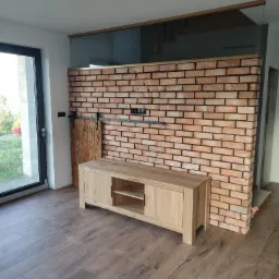 Murowana ściana z czerwonej cegły z czarną fugą, zwieńczona szkłem, drewniana szafka RTV i przesuwne drzwi z surowego drewna na tle podłogi imitującej drewno.