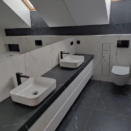 Nowoczesna łazienka na poddaszu z dwoma białymi umywalkami nablatowymi z czarną armaturą, czarnym blatem imitującym marmur, białymi szafkami pod umywalkami, toaletą w zabudowie oraz czarno-białymi...