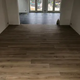 Ułożona podłoga z płytek imitujących drewno, przechodząca w ciemne płytki gresowe w głębi pomieszczenia, z widocznymi krzyżykami dystansowymi i workiem zaprawy przy ścianie.