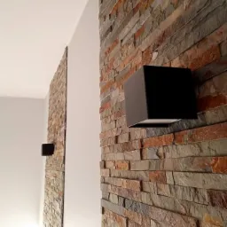 Ściana wyłożona dekoracyjnym kamieniem w odcieniach brązu i szarości, oświetlona dwoma czarnymi, kubicznymi kinkietami.