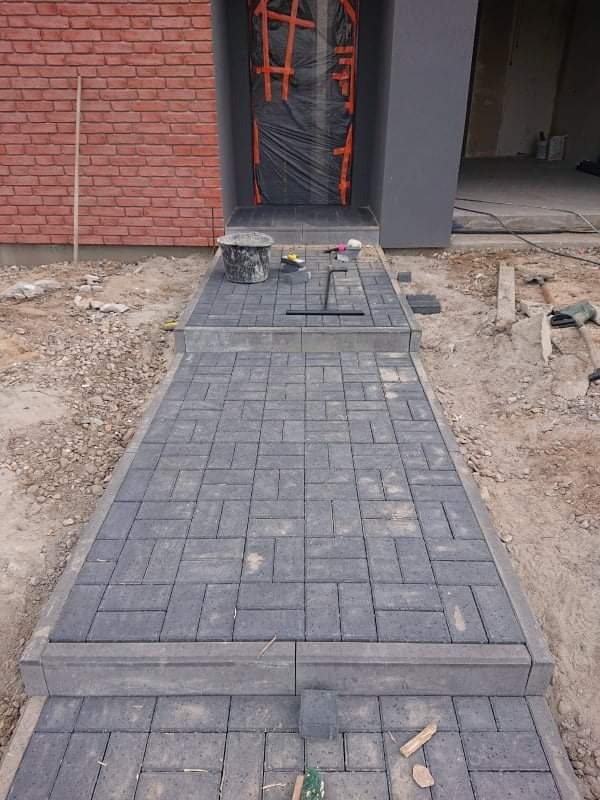 Ułożona ścieżka z szarej kostki brukowej prowadząca do wejścia domu z ceglaną elewacją, widoczne narzędzia i wiadro, drzwi zabezpieczone folią.