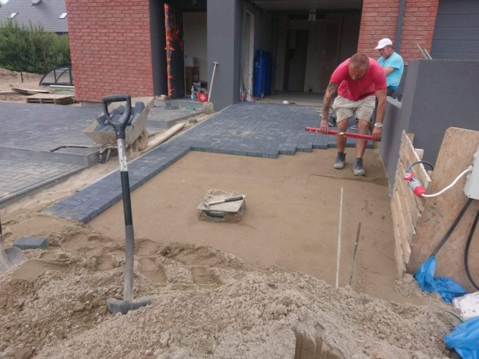 Prace brukarskie w toku: robotnik z poziomicą kontroluje równość ułożonej kostki brukowej na podsypce piaskowej, w tle widoczny drugi pracownik oraz budynek z cegły.