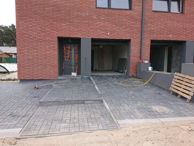 Ułożona szara kostka brukowa przed nowoczesnym domem z cegły, widoczne schody, paleta drewniana i wąż ogrodowy.