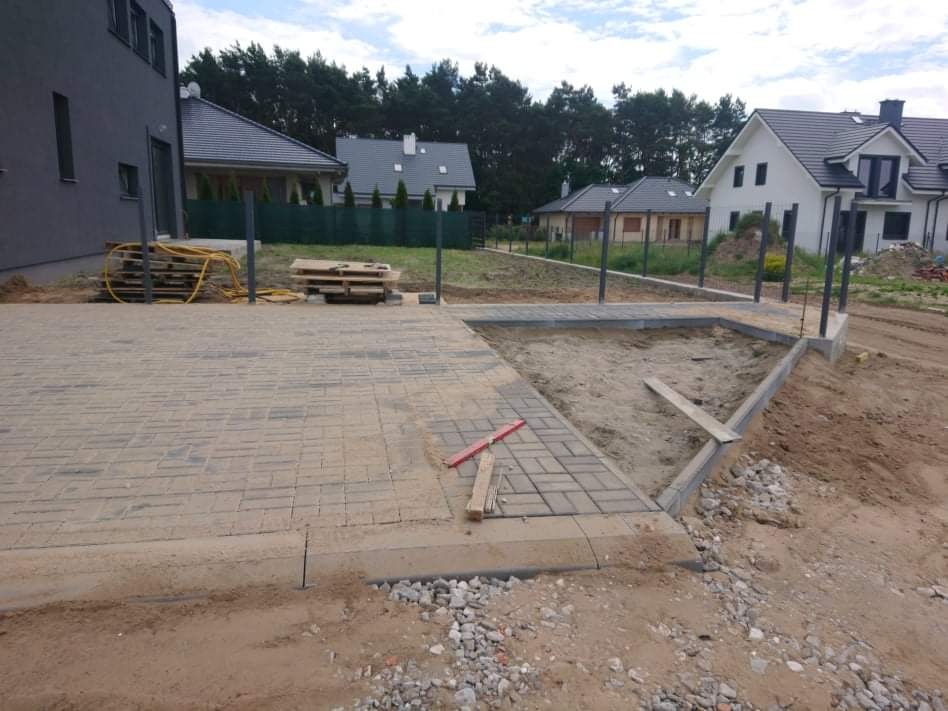 Fragment układanej nawierzchni z kostki brukowej, widoczne obrzeża, piasek, narzędzia, przygotowane podłoże i częściowo ułożona kostka w otoczeniu domów jednorodzinnych.