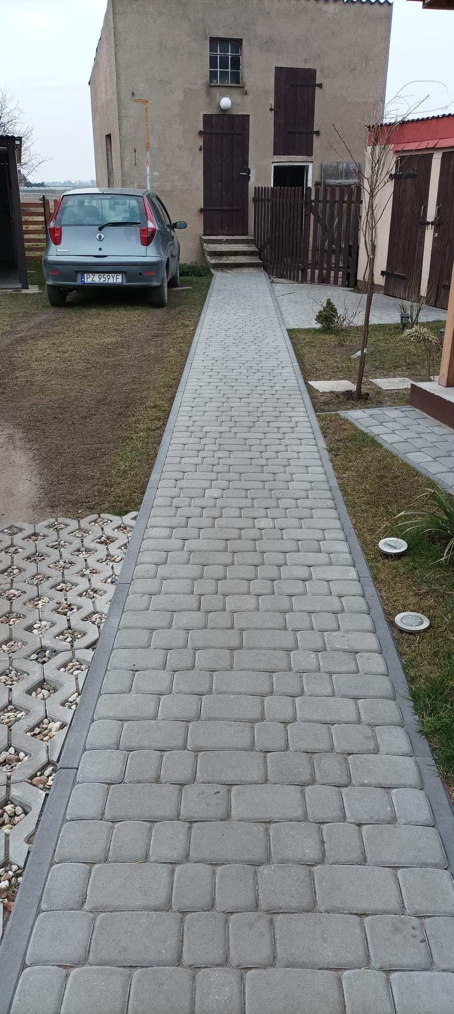 Długa, prosta ścieżka z szarej kostki brukowej prowadząca do budynku z drewnianymi drzwiami i okiennicami, obok zaparkowany szary samochód osobowy.